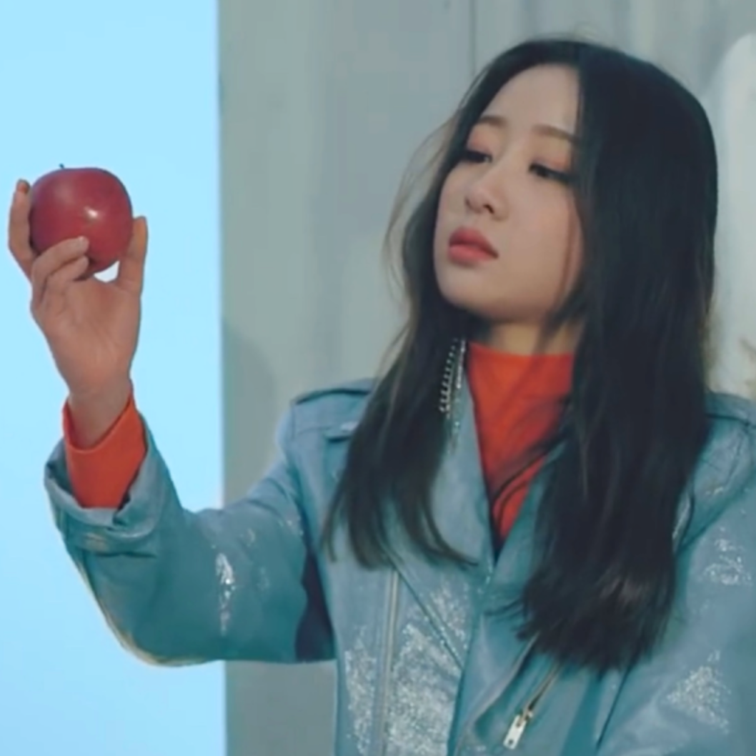 Yves from the 'new' music video. She has long black hair and is wearing a blue jacket. She is holding an apple and gazing at it.